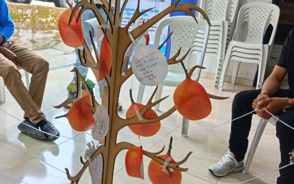 Detalle de una estructura de madera con forma de árbol con ramas que sostienen figuras de cartón que simulan frutos naranjas y tarjetas blancas. En una de las tarjetas se aprecia un mensaje manuscrito dirigido a un ser querido. A los lados se observan personas sentadas en un espacio interior.