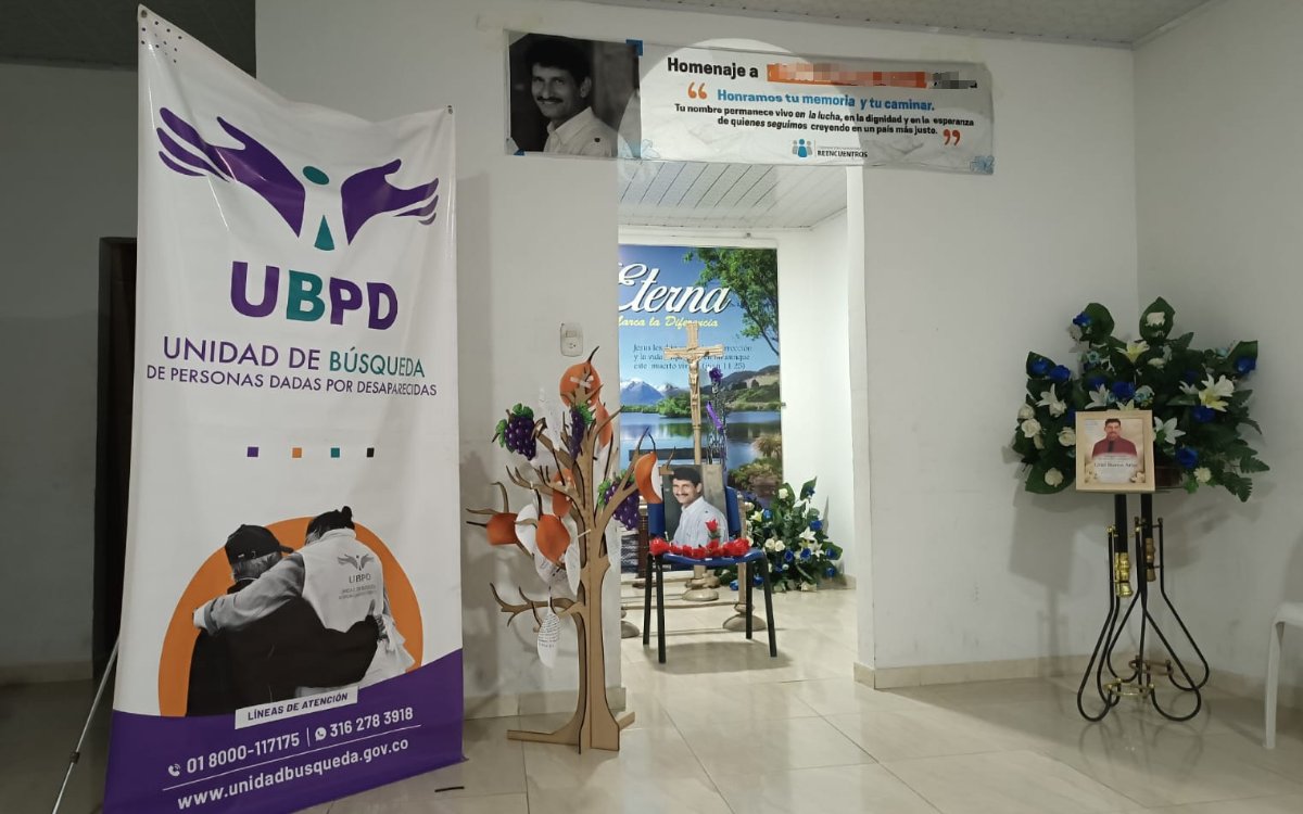 Fotografía de un espacio institucional preparado para un acto solemne. A la izquierda se destaca un pendón con el logo de la Unidad de Búsqueda de Personas dadas por Desaparecidas (UBPD) y sus líneas de atención. En el centro hay un árbol decorativo de madera con mensajes y una pancarta en la pared con el título 