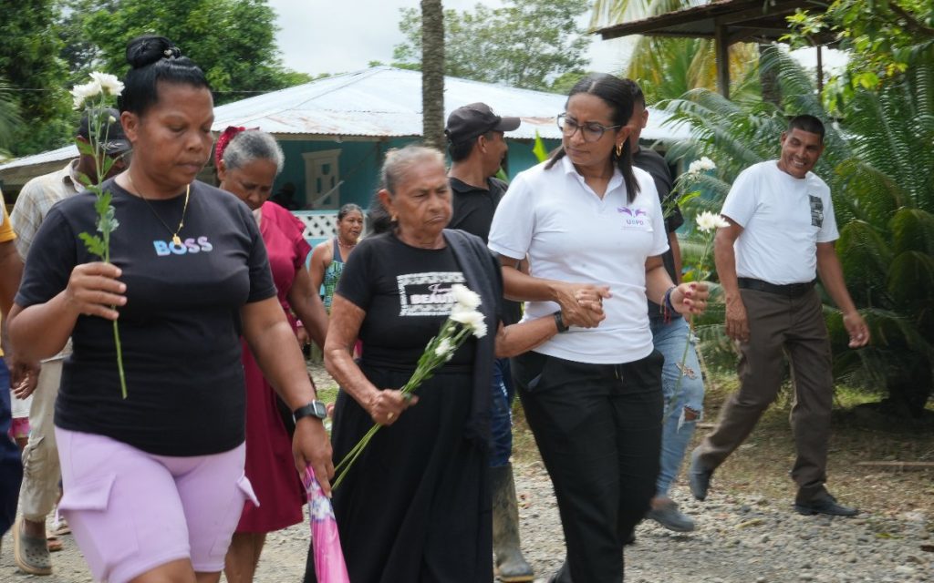 En primer plano, dos mujeres caminan tomadas de la mano: una mujer mayor con vestido negro e insignia en la camiseta, apoyada por una funcionaria con camisa blanca corporativa de la UBPD. A la izquierda, otra mujer afrodescendiente lleva una flor blanca. Al fondo se aprecian viviendas rurales y más allegados acompañando el recorrido.