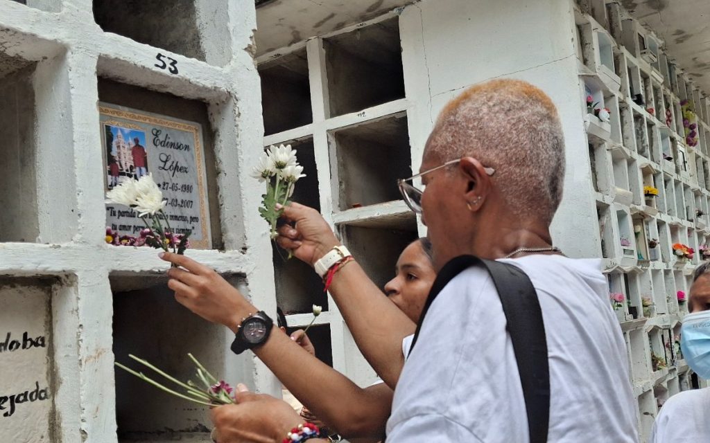 Plano corto en una pared de bóvedas funerarias en un cementerio. Una persona sostiene un ramo de flores blancas y rosas hacia el nicho marcado con la inscripción «Edinson López *27-05-1980 †7-03-2007» junto a la frase «Tu partida dejó un espacio inmenso, pero tu amor permanecerá siempre».
