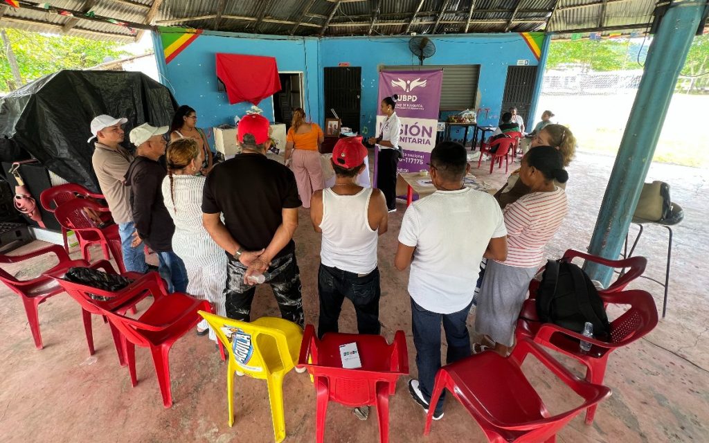 Toma en ángulo picado dentro de un quiosco o caseta comunitaria con techo de lámina. Alrededor de sillas plásticas rojas y amarillas, un grupo de hombres y mujeres escuchan a una representante de la Unidad de Búsqueda que está de pie frente a un pendón institucional con la frase «Misión Humanitaria».