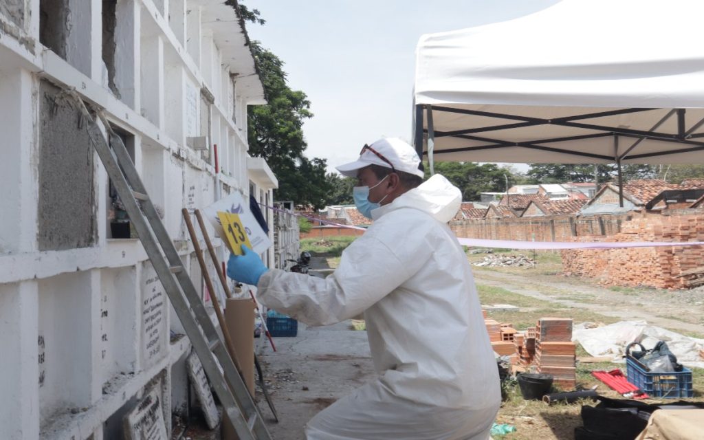 Especialista forense en traje blanco de bioseguridad, guantes y tapabocas coloca un marcador de evidencia amarillo con el número 13 en la lápida de una bóveda de cementerio. A su lado hay una escalera metálica apoyada contra el muro.