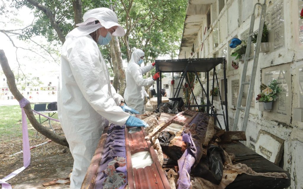 Dos investigadores con trajes blancos de bioseguridad total inspeccionan los restos deteriorados de un ataúd de madera que conserva partes de su forro de tela morada. El cofre reposa sobre una mesa negra frente al pabellón de bóvedas.