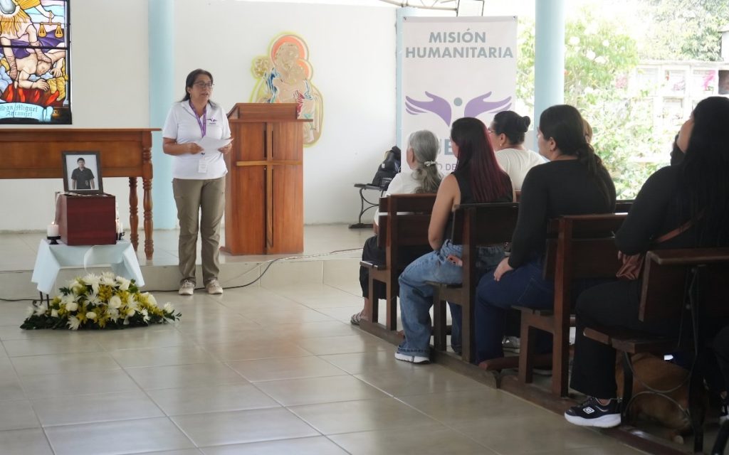 Una mujer con gafas y camiseta blanca con el logo institucional se encuentra de pie leyendo unos papeles en un recinto o capilla. A su izquierda hay un pequeño cofre fúnebre de madera sobre una mesa con velas, flores blancas y la fotografía de un hombre joven. Al fondo, a la derecha, varios asistentes permanecen sentados en bancas escuchando la intervención, al lado de un pendón que dice «MISIÓN HUMANITARIA». En la pared del fondo destaca un vitral con la imagen de San Miguel Arcángel.   