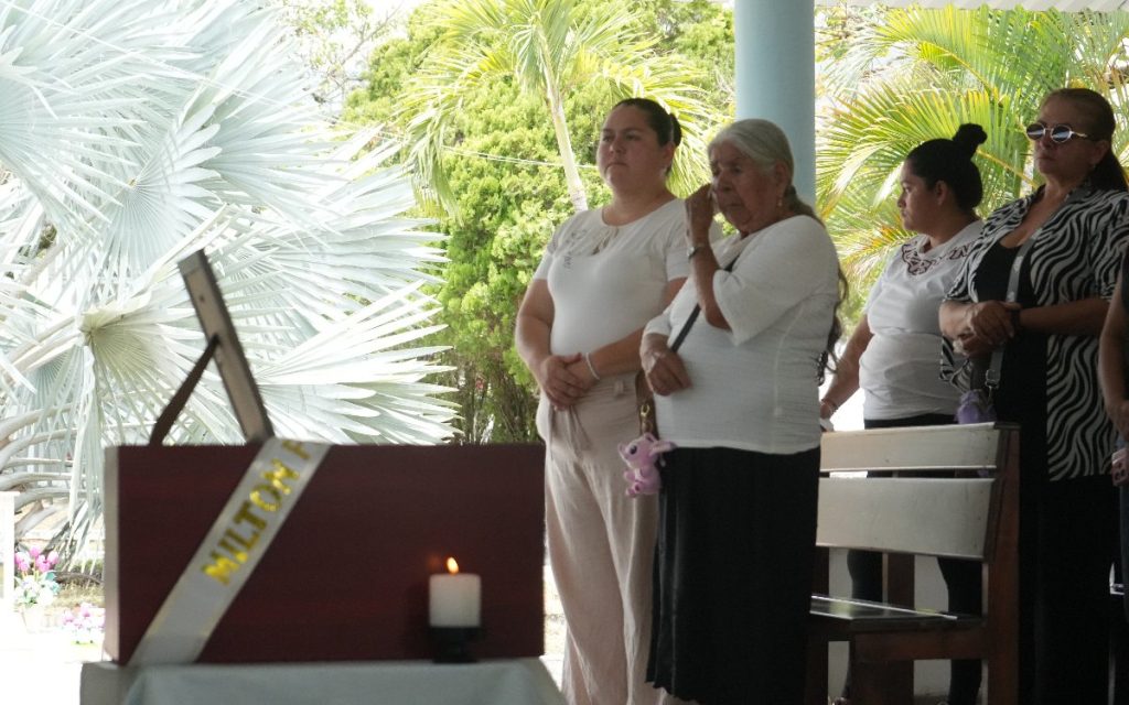 Plano medio en ángulo contrapicado de un grupo de familiares reunidos en un espacio abierto rodeado de palmeras. Una mujer mayor en primer plano limpia sus lágrimas mientras mira hacia el cofre fúnebre de madera, el cual tiene una cinta con el nombre "MILTON" grabado en letras doradas y una vela encendida al lado. A su alrededor, otras personas acompañan en silencio con gestos de emotividad y respeto.