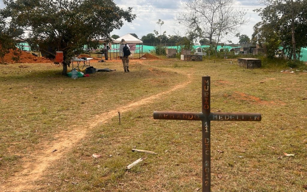 En primer plano se observa una cruz de madera hincada sobre un terreno de césped seco y tierra rojiza. La cruz tiene grabado verticalmente el texto "MURIO" y "11 DEC 2012", e horizontalmente el nombre "ARMANDO MEDELLIN". En el fondo, en una zona despejada del cementerio, hay carpas blancas de trabajo, mesas con equipos y personas realizando labores de campo en la zona rural.