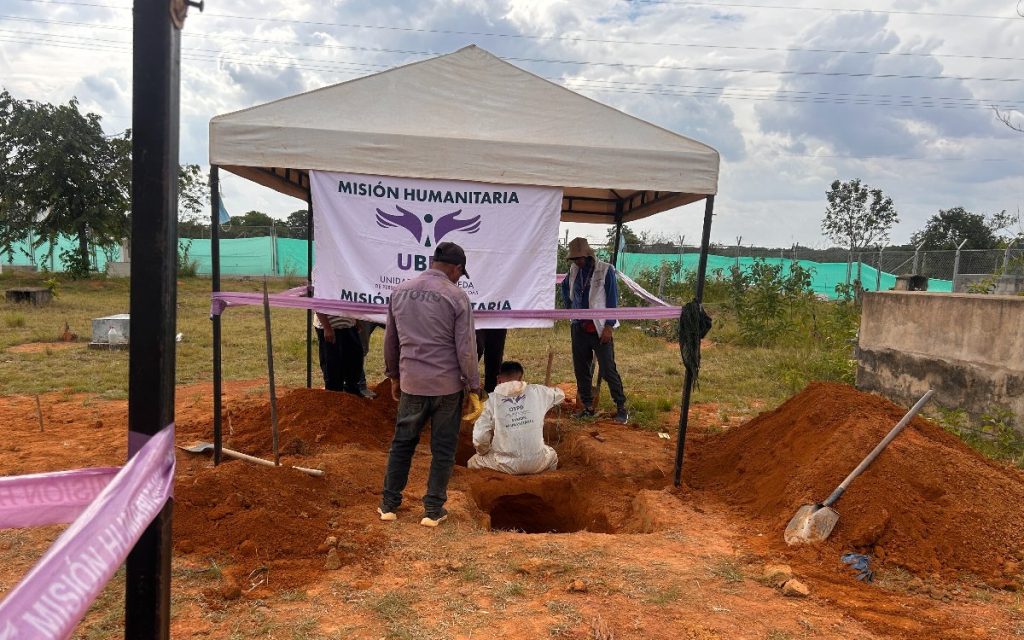Una carpa blanca instalada en un terreno de tierra rojiza cubre una excavación en un cementerio. Frente a la fosa, una persona con traje protector blanco trabaja en su interior, asistida por profesionales de la Unidad de Búsqueda de Personas dadas por Desaparecidas (UBPD). Delante de la carpa se observa una cinta morada con la leyenda "MISIÓN HUMANITARIA".