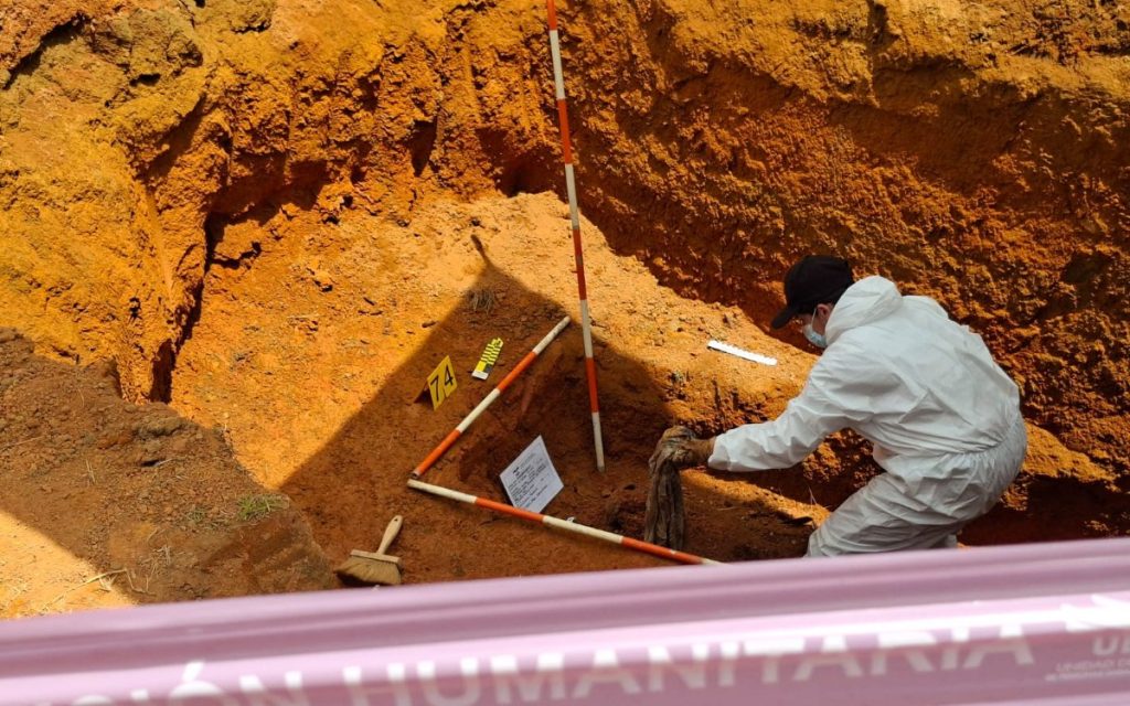 Plano picado del interior de una fosa en terreno rojizo. Un profesional forense vestido con traje biocontaminante blanco y mascarilla manipula evidencia con cuidado. En el suelo de la excavación se aprecian balizas de señalización numeradas con el número 74, regletas de medición forense y herramientas de precisión.