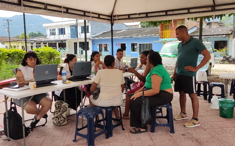 "Fotografía tomada al aire libre bajo una carpa blanca instalada en una plaza pavimentada. Varias personas están sentadas alrededor de mesas portátiles plegables trabajando o conversando frente a computadoras portátiles abiertas de la marca HP. En el costado izquierdo, una mujer con blusa blanca y cabello oscuro usa un portátil; a su lado, otra mujer atiende a otra persona sentada. Al fondo, a la derecha, un hombre de pie con camisa verde observa la escena. Detrás del toldo se aprecian edificios coloridos de un vecindario, vegetación y montañas bajo el cielo despejado."