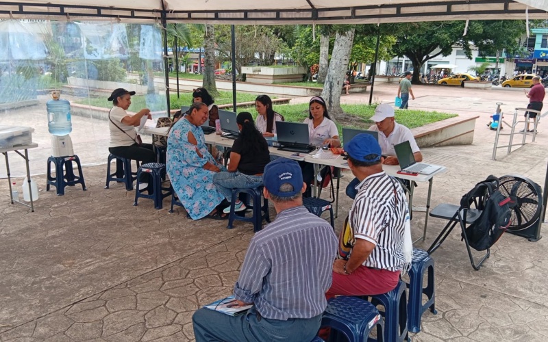 "Fotografía de plano general tomada en una plaza pública pavimentada, donde se ha instalado una carpa blanca grande. Debajo de la estructura, varias personas están sentadas en taburetes plásticos azules alrededor de mesas portátiles largas. En la fila de atención, varios funcionarios operan computadoras portátiles HP atendiendo a los ciudadanos, entre ellos una mujer con blusa azul estampada y otras personas sentadas de frente. En primer plano, de espaldas, se aprecian dos usuarios sentados esperando su turno (un hombre con camisa de rayas y gorra azul, y otro con camisa gris). Al fondo se observa un área abierta con árboles, vehículos transitando en una calle y edificaciones urbanas."