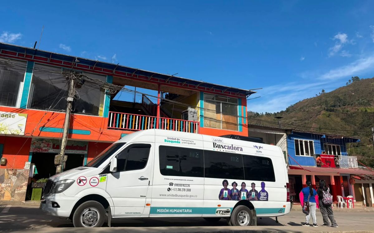 Vista lateral de una camioneta tipo van blanca de la UBPD con los rótulos «Unidad de Búsqueda humanitaria y extrajudicial», «La ruta Buscadora» y «Misión Humanitaria». En su costado lleva un diseño con siluetas de personas sosteniendo retratos, números de contacto e íconos de prohibición de armas. Dos personas caminan junto al vehículo en un entorno rural con edificaciones de fachada naranja y azul, con una montaña al fondo bajo un cielo despejado.