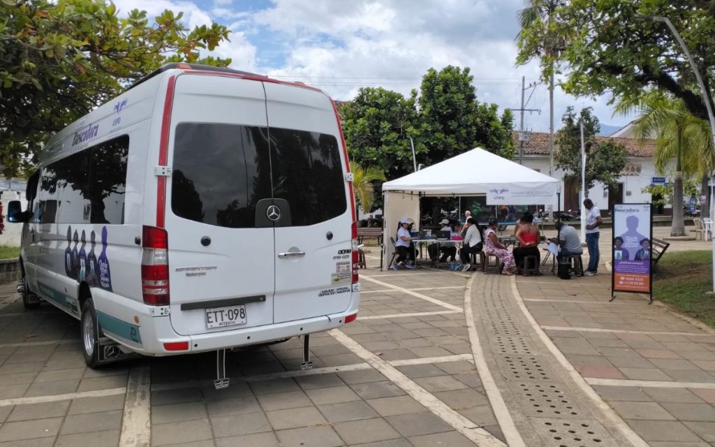 Vista trasera de una camioneta van blanca con placas ETT-098 estacionada sobre el piso adoquinado de una plaza. A un lado, bajo una carpa blanca institucional, varias personas y funcionarios se encuentran sentados atendiendo a los ciudadanos. Junto a la carpa se observa un pendón publicitario con el logotipo de la UBPD y «La ruta Buscadora».