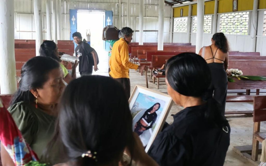 Interior de una iglesia o salón comunitario con paredes de madera y bancos rústicos, donde varias personas se agrupan. En el primer plano, de espaldas, una mujer sostiene un portarretrato con la fotografía de una joven de cabello oscuro. A su izquierda, otra mujer observa con atención, mientras en el fondo, un hombre con camisa amarilla gesticula y un joven carga un pequeño féretro o urna de madera sobre su hombro cerca de la puerta abierta de color azul. Al fondo, se aprecian personas sentadas en las bancas y una mujer de espaldas cerca de otro arreglo floral.
