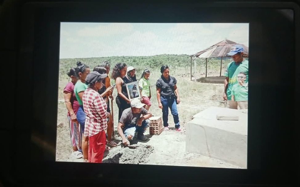 Un grupo de aproximadamente diez personas reunidas al aire libre, en un terreno rural y seco con vegetación y una estructura con techo de zinc al fondo. En el primer plano, un hombre en cuclillas con gorra y ropa informal trabaja en el suelo con una paleta de construcción cerca de una base de concreto o bloque cementado. Detrás de él, varias mujeres y hombres observan con atención; una de las mujeres sostiene un portarretrato con una fotografía. A la derecha, un joven con gorra azul y camiseta verde con un estampado de un rostro observa la escena.