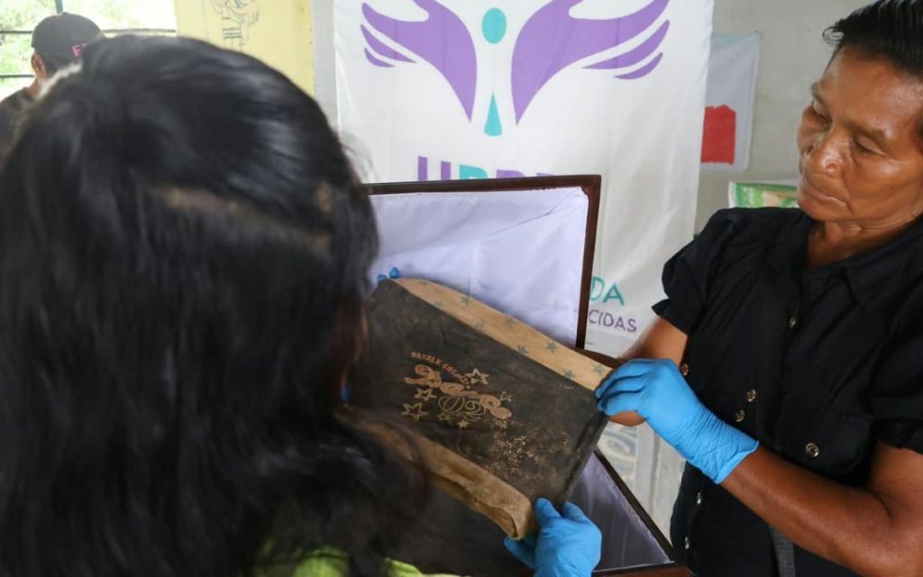 Primer plano de una mujer de cabello oscuro y camisa negra que, usando guantes azules de protección, sostiene y observa con atención un fragmento de tela o prenda antigua, de color oscuro y desgastado, con un estampado que incluye estrellas y la palabra "Danz". Otra persona, de espaldas y con camiseta verde, ayuda a sostener el objeto que está dentro de un cofre o urna de madera con forro interior blanco. Al fondo, se aprecia parte de un pendón blanco con el logotipo de la UBPD y la palabra "...DAS" visible.