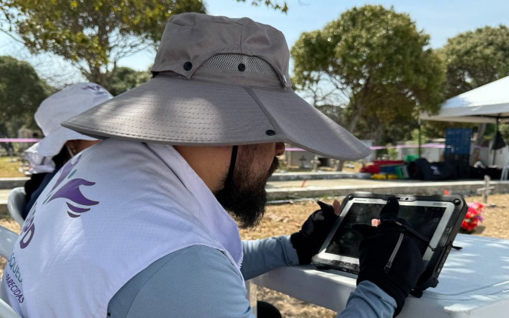 Plano medio lateral de un hombre con sombrero de ala ancha color gris y chaleco blanco institucional con el logo de la UBPD. Utiliza guantes negros y opera una tableta robusta tipo Panasonic sobre una mesa blanca en una jornada de trabajo de campo.
