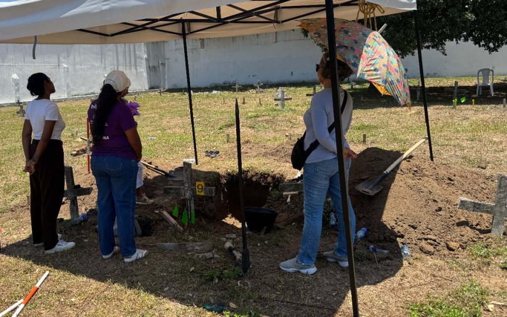 Bajo la sombra de una carpa blanca, tres mujeres observan una fosa de excavación delimitada en un cementerio. Una de ellas sostiene una sombrilla de colores. Junto al pozo se observa tierra removida, palas y cruces de madera sobre el césped.