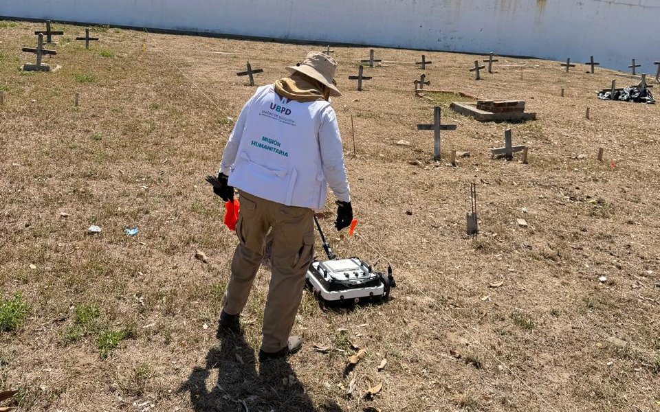 Vista trasera de un especialista de la Misión Humanitaria de la UBPD, vistiendo chaleco blanco identificativo y sombrero de campo. Se encuentra desplazando un equipo de georradar con ruedas sobre el suelo seco de un cementerio lleno de cruces de madera.