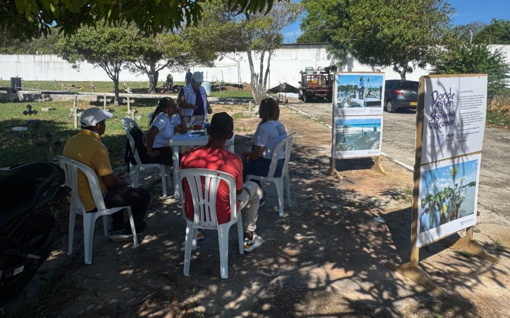 Un grupo de personas y funcionarios de la UBPD sentados en sillas plásticas blancas conversan alrededor de mesas bajo la sombra de un árbol. En primer plano destacan dos paneles informativos de madera con fotografías y el lema "Dónde estén, les seguiremos buscando". Al fondo se aprecia un cementerio y un vehículo de trabajo.
