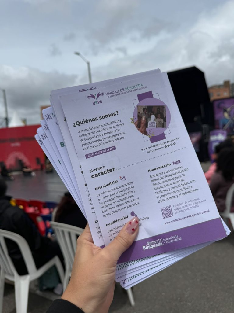 Una mano sostiene varios volantes impresos. En la portada del primer folleto se aprecia el logotipo de la UBPD con el texto "¿Quiénes somos?" y secciones sobre su carácter extrajudicial, confidencial y humanitario, con un código QR y enlace web en la parte inferior.