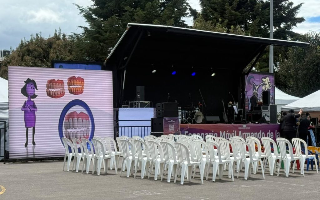 Vista frontal de una plaza o evento al aire libre con filas de sillas plásticas blancas y de colores vacías frente a un escenario móvil techado. A la izquierda, una gran pantalla electrónica exhibe la figura animada de un personaje violeta junto a diagramas de dentadura.
