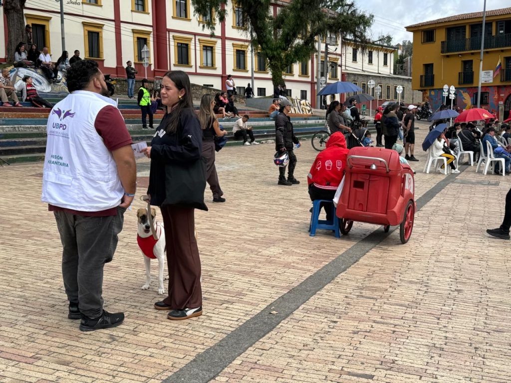 Fotografía en plano medio tomada en una plaza de adoquines. En primer plano a la izquierda, un hombre de espaldas porta un chaleco blanco de la UBPD con el logo institucional y la leyenda "Misión Humanitaria". Habla con una mujer vestida con ropa oscura que sostiene la correa de un perro pequeño con pechera roja. Al fondo se observan graderías con personas sentadas, un vendedor ambulante sentado junto a un carrito rojo de helados y edificaciones tradicionales de fondo.