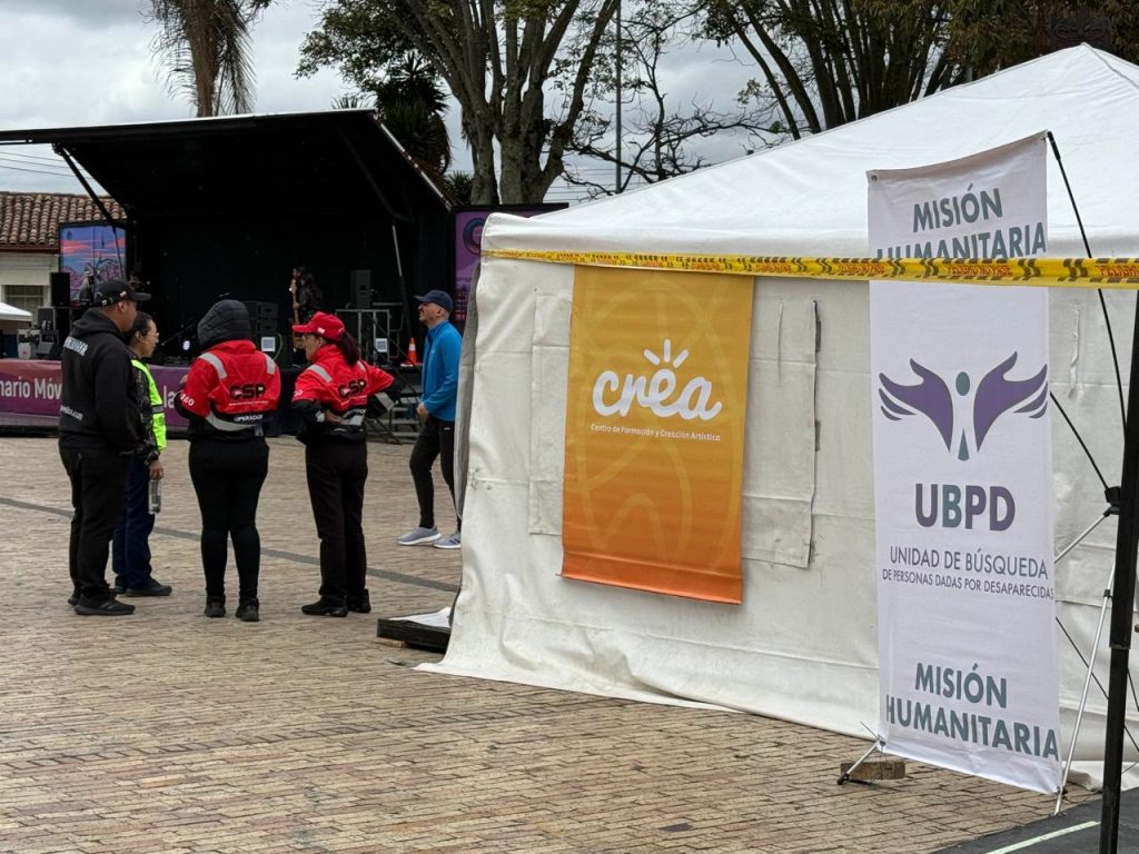 En primer plano a la derecha se observa un pendón vertical blanco con el logotipo violeta de la UBPD y las palabras "UBPD - Unidad de Búsqueda de Personas dadas por Desaparecidas" y "Misión Humanitaria". Detrás, una carpa blanca tiene colgado un pendón naranja del programa "Crea - Centro de Formación y Creación Artística". A la izquierda, un grupo de cuatro personas de logística con chaquetas rojas y gorras conversan frente a un escenario al aire libre.
