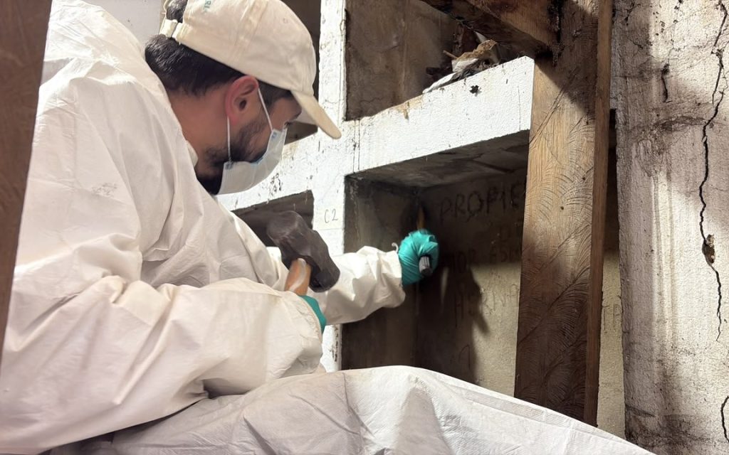 Un integrante del equipo forense con overol blanco, mascarilla, gorra y guantes utiliza una mandarria pequeña para abrir o inspeccionar una bóveda fúnebre con marcas en la superficie.