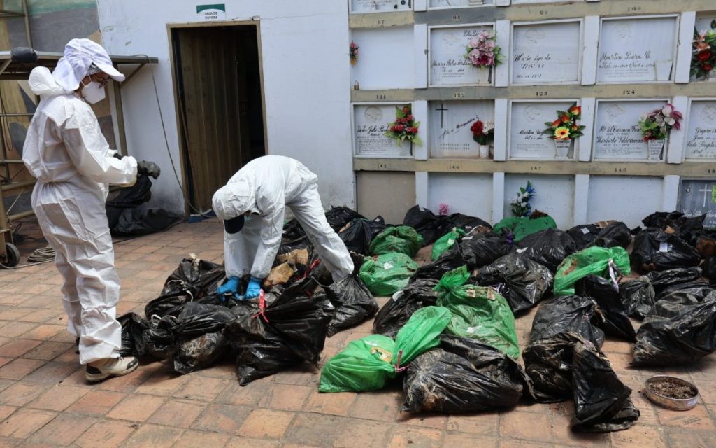 En el patio del cementerio, rodeado por paredes de osarios y nichos con flores, dos personas del equipo forense clasifican y revisan en el suelo numerosas bolsas negras y verdes con material recuperado.