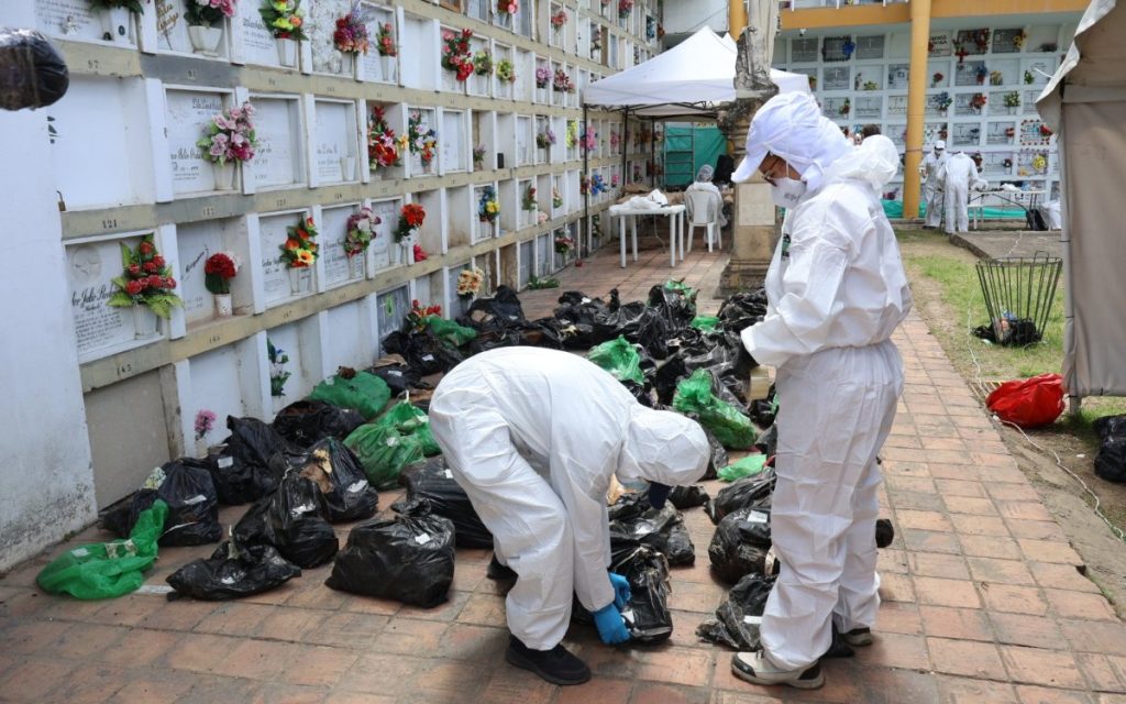 Vista panorámica del pasillo del cementerio donde dos miembros del equipo con indumentaria completa de bioseguridad precintan y ordenan bolsas de recuperación alineadas en el suelo de baldosas.