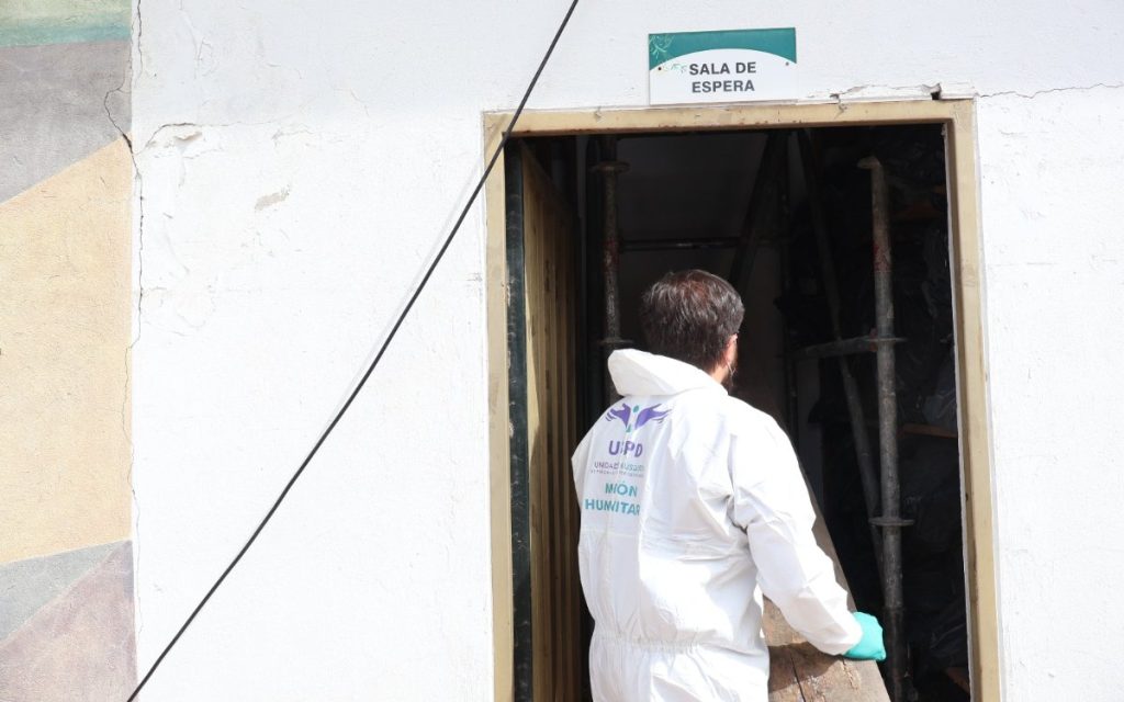 Vista de espaldas de un trabajador que lleva en su overol el logo de la Unidad de Búsqueda de Personas Dadas por Desaparecidas (UBPD - Misión Humanitaria), entrando por una puerta con el cartel «Sala de espera».