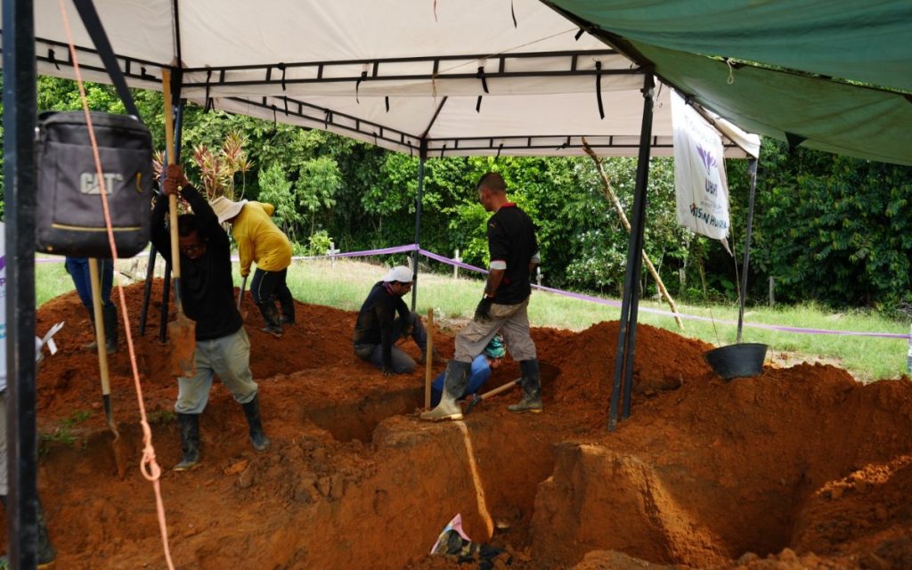 Vista en plano medio debajo de una carpa de lona blanca donde un grupo de operarios realiza labores de excavación manual en tierra arcillosa de color rojizo. Un hombre usa una pala, otros permanecen agachados en las fosas excavadas y al fondo cuelga un pendón institucional con el logo de la UBPD.