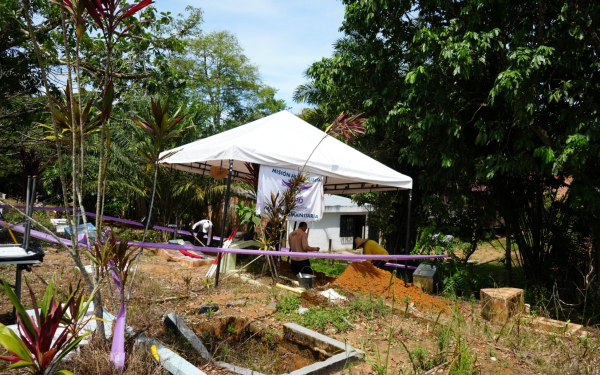 Panorámica al aire libre rodeada de plantas tropicales y árboles. En el centro destaca una carpa blanca de trabajo con un pendón institucional que lleva la leyenda «Misión Humanitaria» y los distintivos de la Unidad de Búsqueda de Personas dadas por Desaparecidas (UBPD). Varios trabajadores realizan labores en el terreno.