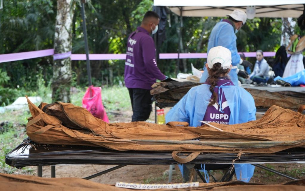 Vista posterior de integrantes del equipo humanitario vistiendo delantales de protección azules y prendas moradas con la sigla «UBPD» grabada en la espalda. Se encuentran trabajando concentrados frente a una mesa de examen dispuesta bajo la carpa de campo.  