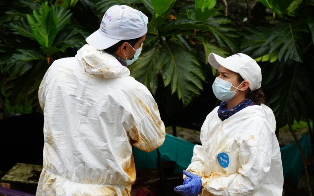 Fotografía en plano medio de dos integrantes del equipo técnico utilizando trajes de bioseguridad tipo Tyvek de color blanco manchados de tierra, gorras blancas institucionales de la UBPD y tapabocas. Ambos profesionales se encuentran cara a cara dialogando en un entorno natural rodeado de vegetación verde.