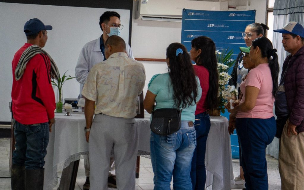 Un grupo de hombres y mujeres vistos desde atrás y de perfil se encuentra reunido en torno a una mesa cubierta con un mantel blanco. En la mesa hay una vela encendida y un florero. Un hombre con bata blanca de profesional de la salud y tapabocas se dirige al grupo. Al fondo, se observa un pendón azul con el logotipo institucional de la JEP (Jurisdicción Especial para la Paz).