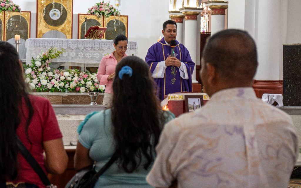 Perspectiva desde la parte trasera de tres asistentes a una misa dentro de una iglesia. En el fondo centrado, un sacerdote con vestidura morada sostiene un micrófono mientras habla frente al altar decorado con flores blancas y doradas. Junto a él se encuentra una mujer con camisa rosada y gafas. Delante del altar reposa el cofre fúnebre con la fotografía de la niña.