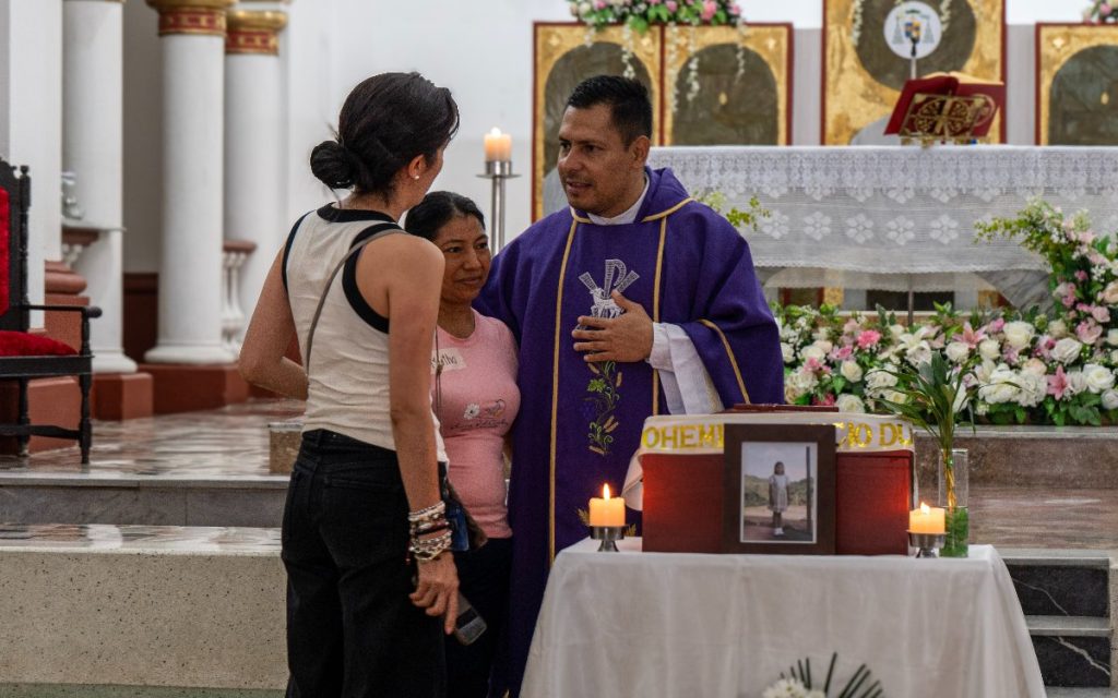 El sacerdote con túnica morada habla de cerca y ofrece consuelo a dos mujeres ubicadas a la izquierda del altar. Una de las mujeres viste una camiseta rosada y la otra una blusa blanca sin mangas. A la derecha en primer plano se observa la mesa con el cofre fúnebre, la foto de la niña y las velas encendidas. El trasfondo muestra el interior iluminado de la iglesia y sus adornos florales.