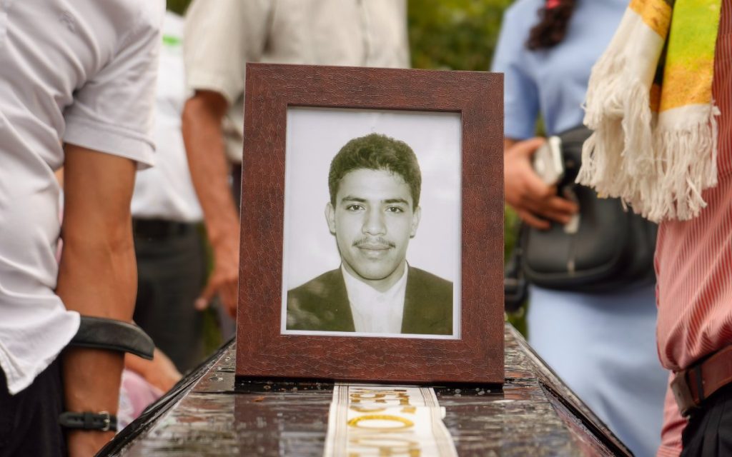 Vista en plano medio de un portarretrato de madera oscura con marco texturizado que contiene la foto en blanco y negro de un hombre joven de cabello corto, tez clara y bigote delgado, vistiendo camisa clara y chaqueta. El portarretrato descansa sobre una superficie cubierta con un plástico o cinta con letras bordadas en dorado, colocada sobre lo que parece ser un féretro. A los lados y al fondo se observan de torso hacia abajo y en plano secundario varias personas vestidas con ropa formal e informal que acompañan el acto.