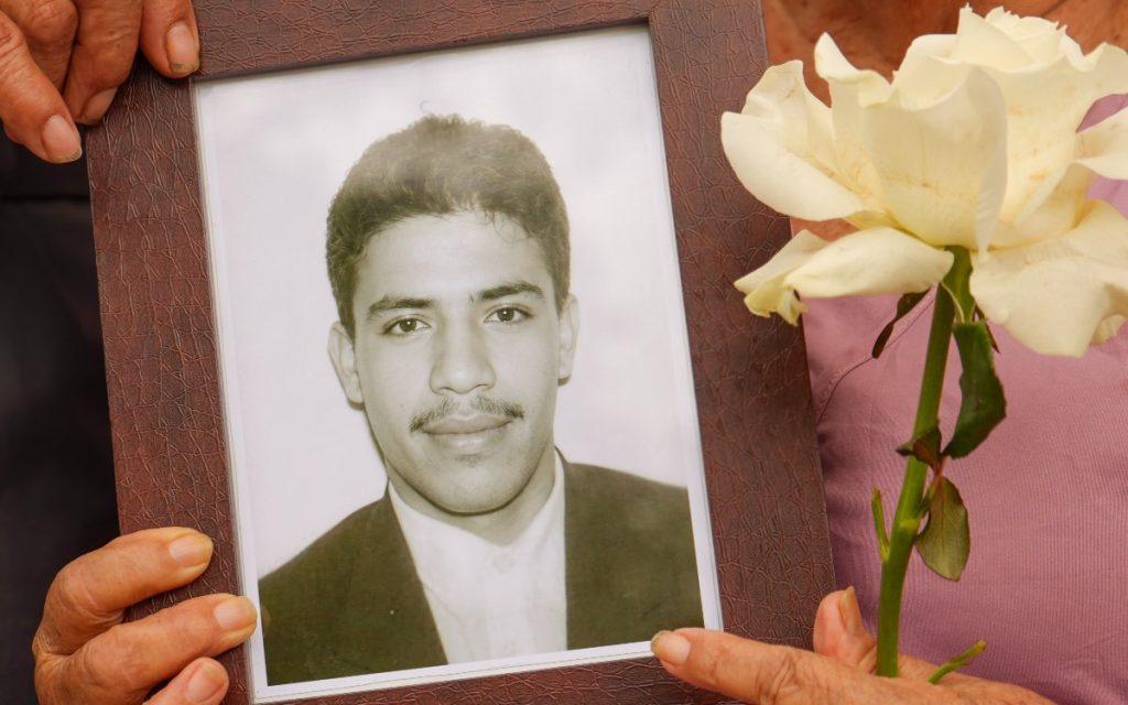 Primer plano en el que dos manos de piel curtida sostienen con firmeza el marco de madera de un retrato en blanco y negro de un hombre joven con bigote. Junto al marco, otra mano sostiene una rosa blanca de tallo verde largo y hoja fresca. La persona que sostiene la flor viste una prenda de tono rosado.