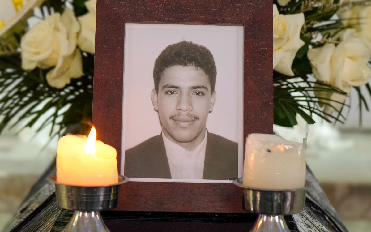 Plano frontal y centrado del portarretrato de madera marrón que contiene la fotografía en blanco y negro del hombre joven. Frente al retrato hay dos candelabros metálicos plateados con velas blancas gruesas: la vela de la izquierda está encendida con una llama viva, mientras que la de la derecha está apagada o baja. Al fondo se observan arreglos florales con rosas blancas.