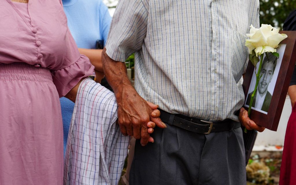 Plano medio corto centrado en los torsos de una pareja. Un hombre vestido con camisa a cuadros de manga corta y pantalón de vestir sostiene con una mano el retrato enmarcado del hombre joven y una rosa blanca. Con su otra mano toma cálidamente la mano de una mujer a su lado, quien viste un vestido rosado con pliegues. Sobre el brazo de la mujer reposa una tela de cuadros tipo pañuelo o paño.