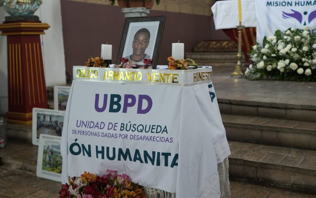 Plano frontal de un cofre ceremonial cubierto por un mantel blanco con el logotipo de la Unidad de Búsqueda de Personas dadas por Desaparecidas (UBPD) y las palabras "Misión Humanitaria". En la parte superior sobresale una cinta con el nombre "Diego Armando Vente" en letras doradas, acompañada por un retrato enmarcado de un joven afrodescendiente con camiseta a rayas, dos velas blancas encendidas y arreglos florales.   