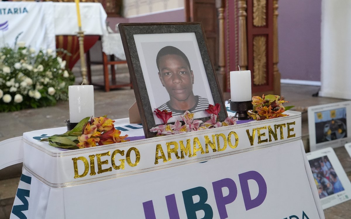 Primer plano en ángulo tres cuartos del retrato enmarcado de Diego Armando Vente. La foto muestra a un joven afrodescendiente con ropa a rayas. Frente al marco reposan flores anaranjadas y rosadas, dos velas blancas y una cinta reflectiva con el nombre 