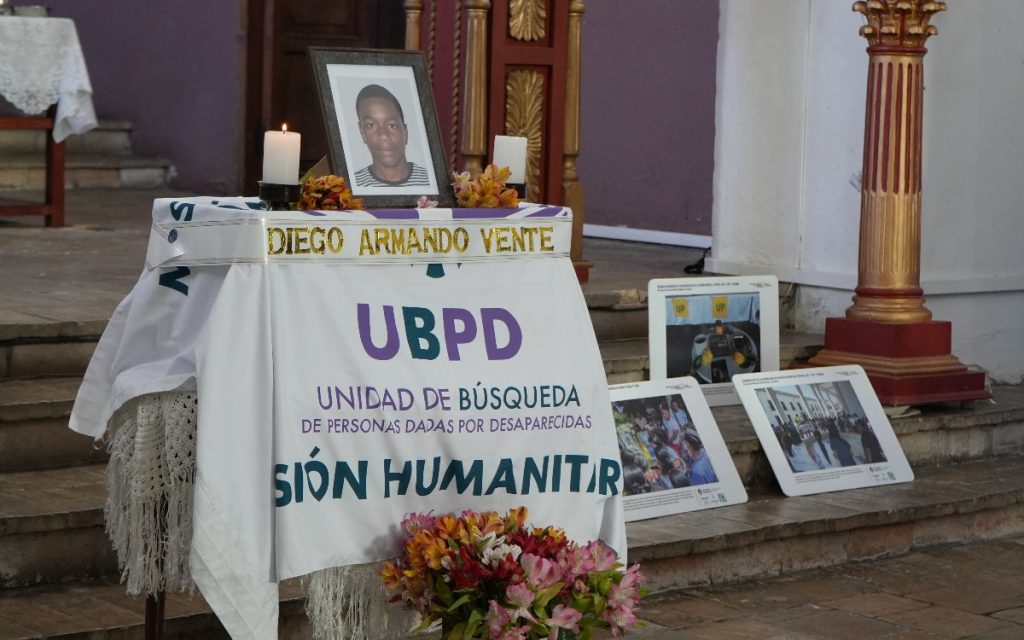 Plano general del altar preparado para la entrega digna. En el centro, sobre unos escalones de piedra, se ubica la estructura cubierta con la tela de la UBPD, el retrato de Diego Armando Vente, velas encendidas y flores. A la derecha, recostados sobre los escalones, se observan tres carteles pedagógicos con fotografías e información del proceso.