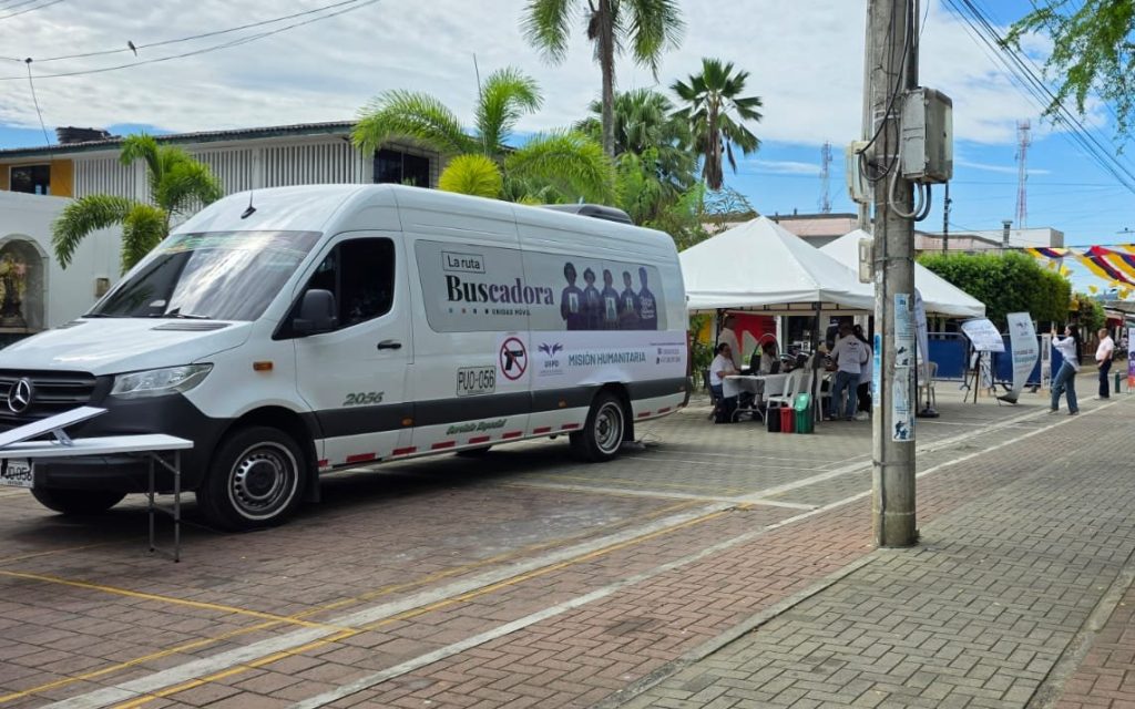 Una camioneta van blanca con los emblemas de la Unidad de Búsqueda de Personas dadas por Desaparecidas (UBPD) y el texto "La Ruta Buscadora - Misión Humanitaria". A su lado hay carpas blancas donde personal institucional atiende al público en un espacio al aire libre con palmeras y edificaciones de fondo.