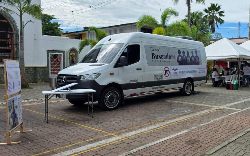 Plano medio de la camioneta van blanca de la UBPD con la inscripción "La Ruta Buscadora - Misión Humanitaria". En primer plano, a la izquierda, hay un marco de madera con carteles informativos del proceso de búsqueda de personas desaparecidas y una mesa plegable.