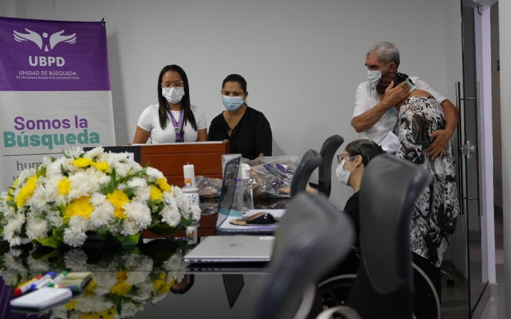 Fotografía de un acto institucional. A la izquierda se observa un pendón morado de la Unidad de Búsqueda de Personas dadas por Desaparecidas (UBPD) con el lema "Somos la Búsqueda". Frente a una mesa decorada con flores amarillas y blancas, dos profesionales con tapabocas atienden la entrega. A la derecha, un hombre mayor abraza a una mujer visiblemente emocionada para brindarle consuelo.