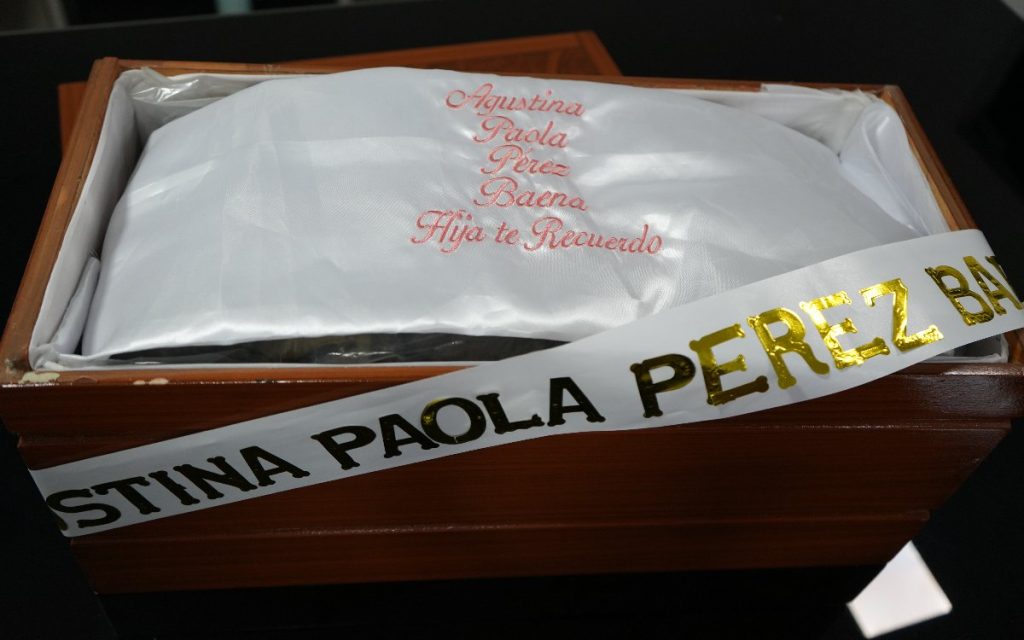 Primer plano superior de una caja o cofre funerario de madera. En su interior hay una funda de satén blanco con una dedicatoria bordada en hilo rosado que dice: "Agustina Paola Pérez Baena / Hija te Recuerdo". Cruzando la caja se aprecia una cinta blanca con letras doradas que lleva el nombre completo de la persona.