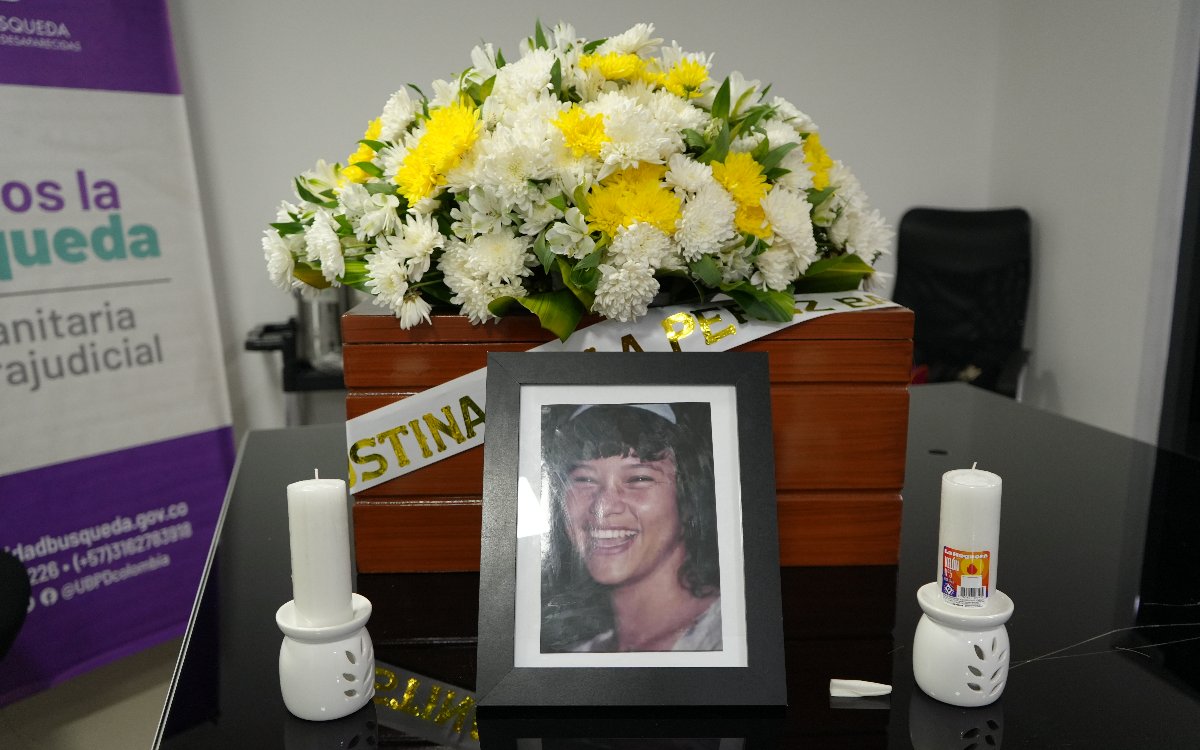 Fotografía centrada en un altar conmemoraivo. En primer plano, un portarretratos negro contiene la fotografía en blanco y negro de Agustina Paola Pérez Baena sonriendo. Flanqueando el retrato hay dos velones blancos encendidos sobre portavelas de cerámica. Al fondo se ubica el cofre de madera cubierto por un gran arreglo de flores blancas y amarillas.
